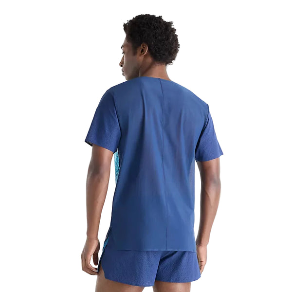 En mand står med ryggen til kameraet og er iklædt et matchende sæt bestående af en blå, kortærmet New Balance Race Day Ultra Light Printed T-shirt, Herre, Altitude og shorts. T-shirten er ikke stoppet ned i bukserne og har en lige nederkant, hvilket afspejler stilen på Race Day Ultra Light Printed T-shirten. Baggrunden er ensfarvet hvid.