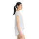 En kvinde med hestehale står i profil iført New Balance Race Day Ultra Light T-Shirt til kvinder i Grey Matter og hvide NB-shorts på en almindelig hvid baggrund.