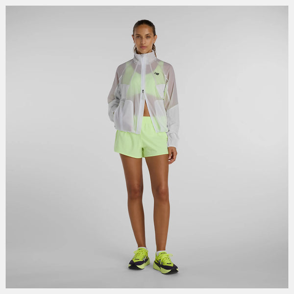 En kvinde står med ansigtet fremad og er iført en New Balance Rc Jacket, Dame, Grey-zip-up over en lysegrøn top og shorts med matchende neongrønne sportssko på en almindelig lysegrå baggrund.