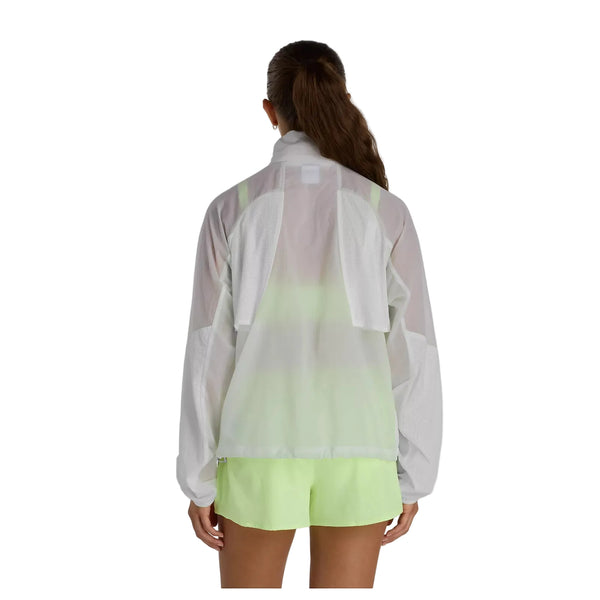 En kvinde står og kigger væk i en New Balance Rc Jacket, Dame, Grey over et neongult-grønt sportssæt med shorts. Hendes brune hår er sat op i en lav hestehale mod en almindelig hvid baggrund.