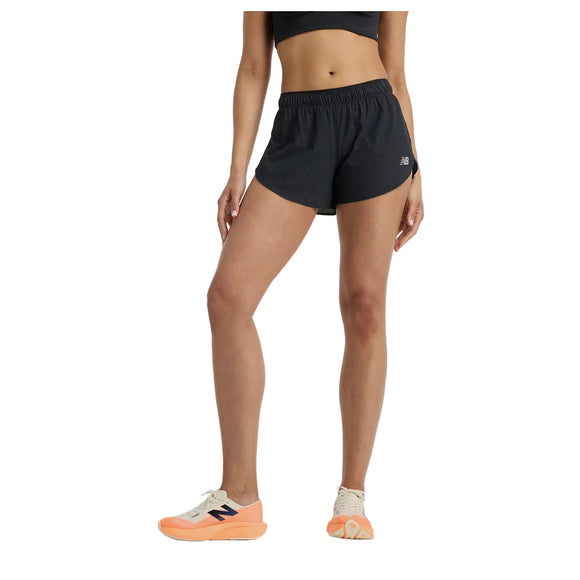 En person står på en hvid baggrund, hvor man kun kan se underkroppen og benene, iført sorte New Balance RC Ultra Light Split Short 3" dameshorts og hvide sneakers med orange såler.