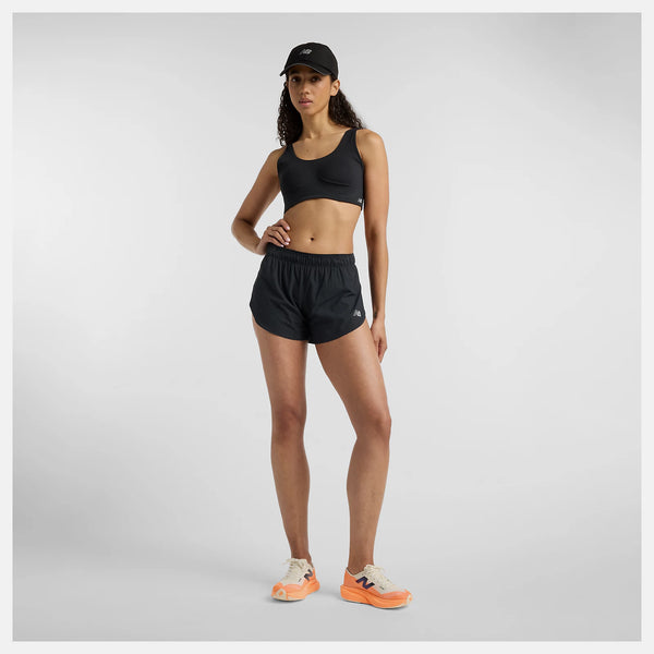 En kvinde vender sig mod kameraet med en hånd på hoften iført en sort sports-bh, New Balance Rc Ultra Light Split Short 3" Dame i sort, en sort kasket og lyse løbesko med orange såler på en ensfarvet baggrund.