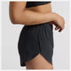 En kvinde ses fra siden i en sort sports-bh og New Balance Rc Ultra Light Split Short 3", Dame, Black med split i siden. Hendes højre arm er bøjet, venstre hånd ligger afslappet ved siden af hende på en almindelig lysegrå baggrund.