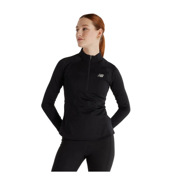 En kvinde står med hænderne bag ryggen og kigger let til siden foran en ensfarvet hvid baggrund, iført den sorte New Balance Rc Essential Heat Grid 1/2 Zip.