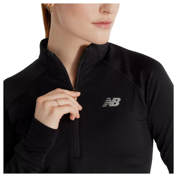 En kvinde iført en sort New Balance Rc Essential Heat Grid 1/2 Zip-løbetrøje med logoet på brystet, der holder fast i lynlåsen med den ene hånd og står let vendt til siden foran en hvid baggrund. Denne alsidige løbetrøje til kvinder er perfekt til løbeture i køligt vejr.
