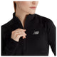 En kvinde iført en sort New Balance Rc Essential Heat Grid 1/2 Zip-løbetrøje med logoet på brystet, der holder fast i lynlåsen med den ene hånd og står let vendt til siden foran en hvid baggrund. Denne alsidige løbetrøje til kvinder er perfekt til løbeture i køligt vejr.