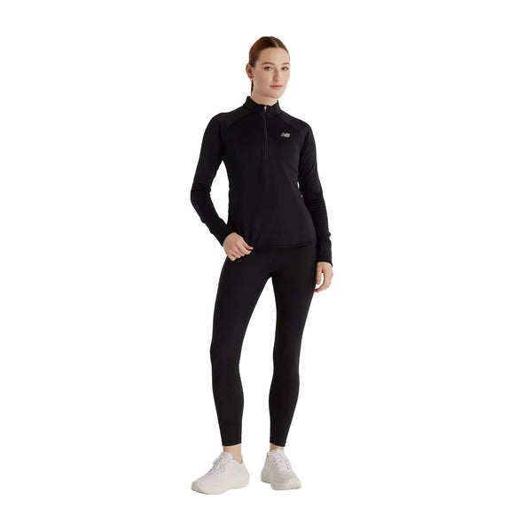 En person står med ansigtet vendt fremad og har en New Balance Rc Essential Heat Grid 1/2 Zip, Dame i sort på sig sammen med sorte leggings og hvide sneakers. Baggrunden er helt hvid.