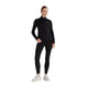 En person står med ansigtet vendt fremad og har en New Balance Rc Essential Heat Grid 1/2 Zip, Dame i sort på sig sammen med sorte leggings og hvide sneakers. Baggrunden er helt hvid.