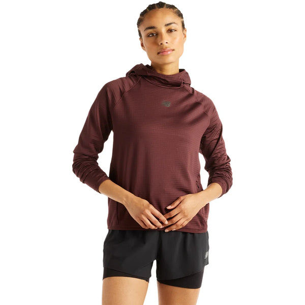 En person står med ansigtet vendt fremad iført New Balance Rc Essential Heat Grid Hoodie, Dame, i farven Cherry Leather med NB HEAT-teknologi og sorte sportsshorts foran en ensfarvet hvid baggrund – perfekt til løb i koldt vejr.