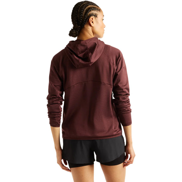Man ser en person med flettet hår bagfra. Personen bærer en rødbrun New Balance Rc Essential Heat Grid-hoodie, Dame, Cherry Leather, udstyret med NB HEAT-teknologi, samt sorte shorts. Personen står foran en ensfarvet hvid baggrund med armene afslappet ned langs siden, klar til at løbe i koldt vejr.