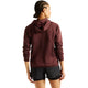 Man ser en person med flettet hår bagfra. Personen bærer en rødbrun New Balance Rc Essential Heat Grid-hoodie, Dame, Cherry Leather, udstyret med NB HEAT-teknologi, samt sorte shorts. Personen står foran en ensfarvet hvid baggrund med armene afslappet ned langs siden, klar til at løbe i koldt vejr.