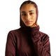En kvinde med flettet hår har New Balance Rc Essential Heat Grid Hoodie i farven Cherry Leather på, der er udstyret med NB HEAT-teknologi, mens hun ser ind i kameraet med et neutralt udtryk og den ene hånd tæt på hagen. Med en ensfarvet hvid baggrund i baggrunden træder dette perfekte mellemlag til løb i køligt vejr frem i forgrunden.