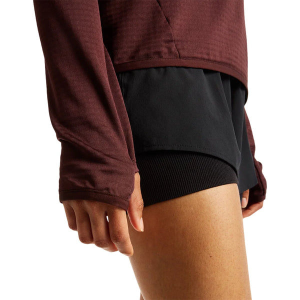 En person iført en langærmet New Balance Rc Essential Heat Grid-hoodie (Dame) i farven »Cherry Leather« og sorte sportsshorts står med venstre arm afslappet ned langs siden. Billedet er beskåret ved det øverste lår og midt på overkroppen.