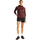 En person står med ansigtet vendt fremad og har en Rc Essential Heat Grid-hoodie til kvinder fra New Balance i kirsebærrødt læder med NB HEAT-teknologi på, sorte sportsshorts, hvide sokker og sorte sneakers. Baggrunden er ensfarvet hvid.