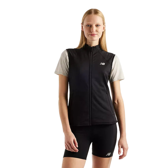 En person iført en lysegrå skjorte med korte ærmer, sorte sportsshorts med New Balance-logoer og en sort New Balance RC Essential Heat Grid-vest til kvinder står foran en ensfarvet hvid baggrund.