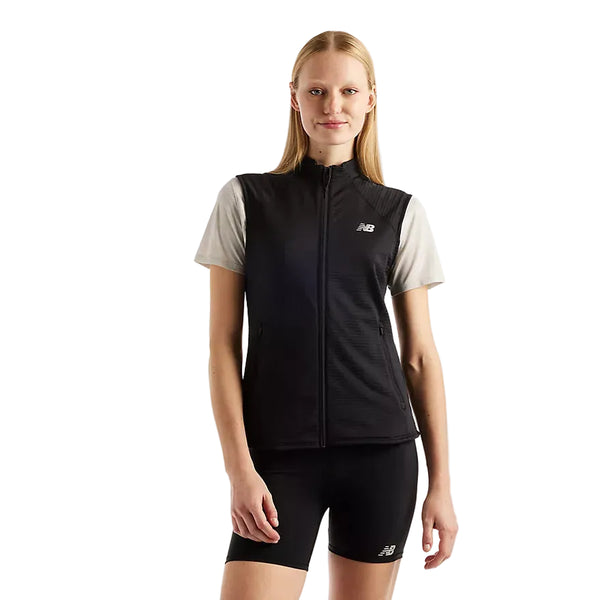 En person iført en lysegrå skjorte med korte ærmer, sorte sportsshorts med New Balance-logoer og en sort New Balance RC Essential Heat Grid-vest til kvinder står foran en ensfarvet hvid baggrund.
