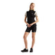 En person står foran en hvid baggrund iført en sort New Balance RC Essential Heat Grid Vest (Dame) over en lys kortærmet skjorte, kombineret med sorte cykelshorts og sorte sportssko. Hænderne hviler let på taljen.