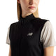 En kvinde med glat, blondt hår har en sort New Balance Rc Essential Heat Grid-vest på over en kortærmet, lysegrå sportstrøje. Billedet er beskåret fra skuldrene og opefter.