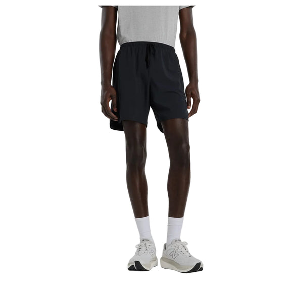 En person iført en grå T-shirt, hvide sokker, hvide sneakers og et par New Balance Rc Essential Short 7" (Herre, sort) står foran en hvid baggrund. Personens hoved er ikke synligt på billedet.