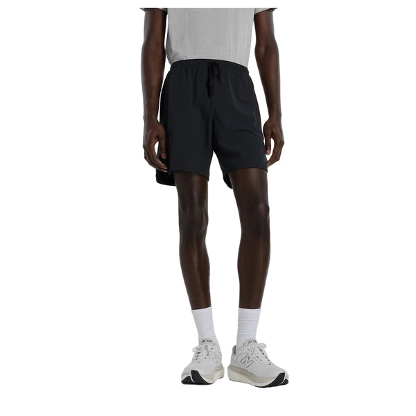 En person iført en grå T-shirt, hvide sokker, hvide sneakers og et par New Balance Rc Essential Short 7" (Herre, sort) står foran en hvid baggrund. Personens hoved er ikke synligt på billedet.