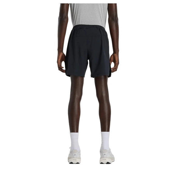 Der ses en person set bagfra på en ensfarvet hvid baggrund, iført en grå T-shirt, et par New Balance Rc Essential Short 7" Herre Black, hvide sokker og hvide sneakers.