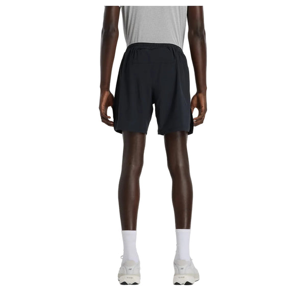 Der ses en person set bagfra på en ensfarvet hvid baggrund, iført en grå T-shirt, et par New Balance Rc Essential Short 7" Herre Black, hvide sokker og hvide sneakers.