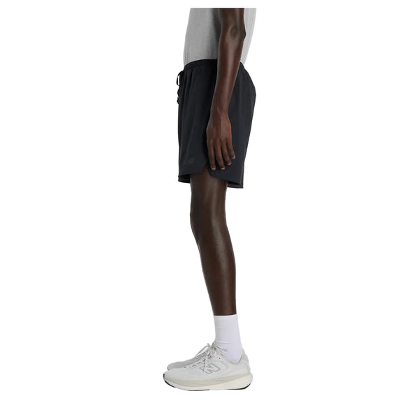 En person set i profil er iført en grå T-shirt, sorte New Balance Rc Essential Short 7" (Herre), hvide ankelstrømper og hvide sportssko foran en ensfarvet hvid baggrund.