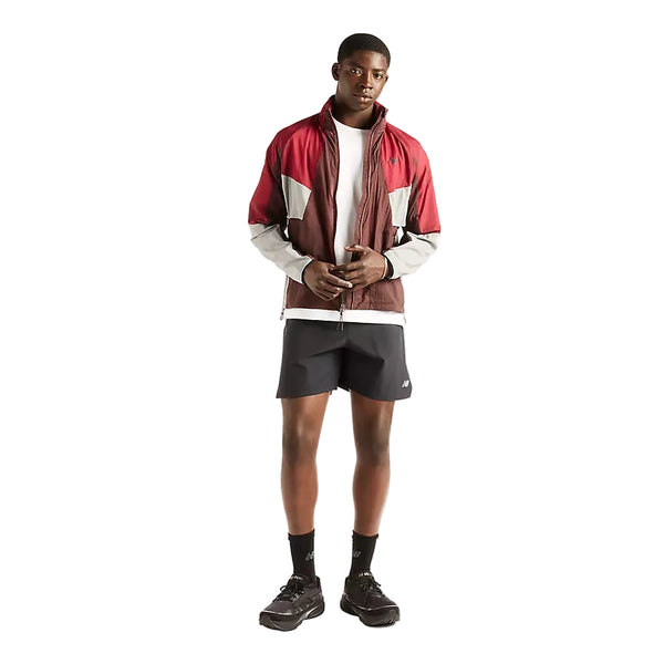 En mand står med ansigtet vendt fremad iført Rc Jacket, Herre, Cherry Leather fra New Balance – en rød og hvid vandafvisende jakke – sammen med en hvid T-shirt, sorte shorts, sorte sokker og sorte sneakers. Han holder hænderne samlet og står foran en ensfarvet hvid baggrund.