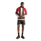 En mand står med ansigtet vendt fremad iført Rc Jacket, Herre, Cherry Leather fra New Balance – en rød og hvid vandafvisende jakke – sammen med en hvid T-shirt, sorte shorts, sorte sokker og sorte sneakers. Han holder hænderne samlet og står foran en ensfarvet hvid baggrund.