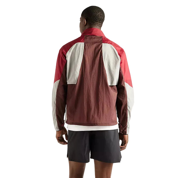 En person står med ryggen til og har den elegante New Balance Rc-jakke til mænd i farven »Cherry Leather« på – en løbejakke i farvekombinationen maroon, rød og grå – samt sorte shorts. Baggrunden er helt hvid.