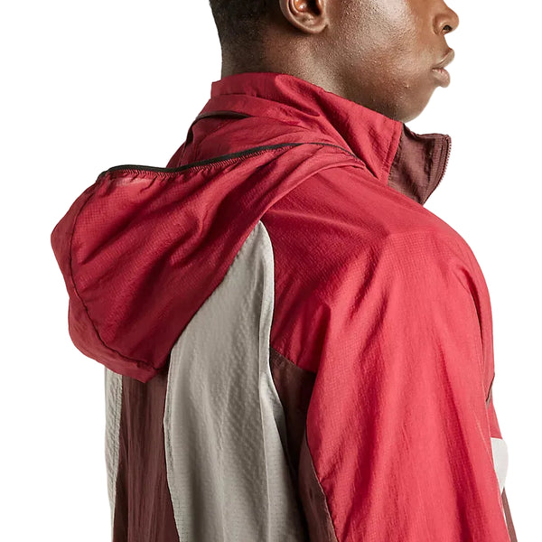 En person har New Balance RC-jakken på (herre, farven Cherry Leather). Billedet viser et sidebillede fra skulderen og opefter, hvor der lægges vægt på jakkens materiale og detaljerne på hætten for at fremhæve dens kvalitet som en vandafvisende jakke. Baggrunden er ensfarvet og diskret.