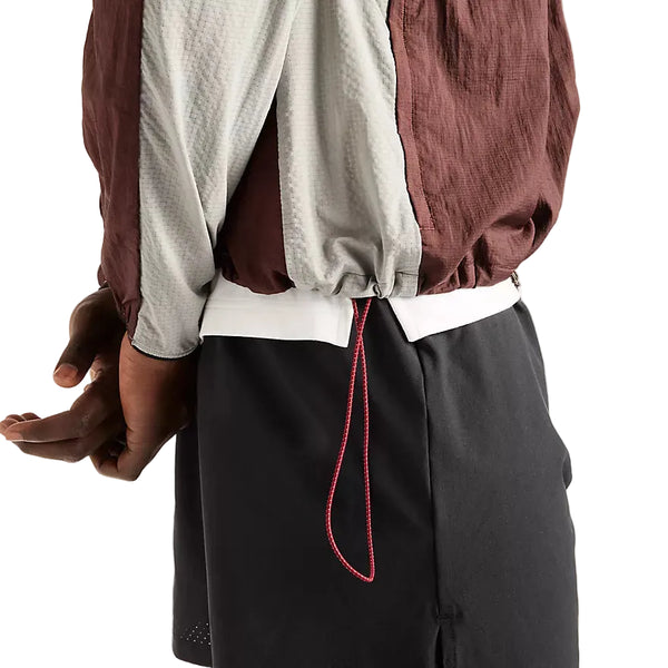 En person står med venstre arm afslappet og er iført en New Balance Rc-jakke, Herre, Cherry Leather, med en synlig rød snor og sorte shorts. På billedet ses kun overkroppen, venstre arm og lårene.