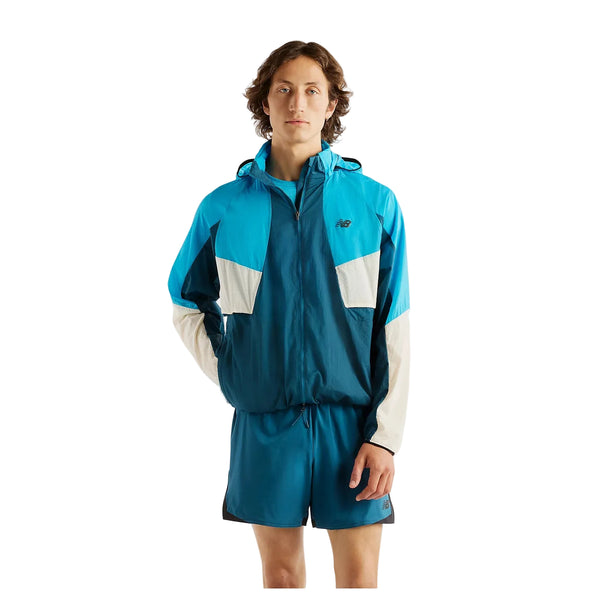 En person står med ansigtet vendt fremad iført en New Balance Rc-jakke, Herre, Streamsong – en let vandafvisende jakke – med hvide detaljer og matchende blå sportsshorts foran en ensfarvet hvid baggrund.