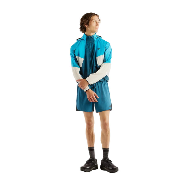 En person med kort brunt hår, der kigger til siden, står foran en hvid baggrund iført en blå og hvid New Balance Rc-jakke (Herre, Streamsong), matchende blå shorts, sorte sokker og sorte sko.