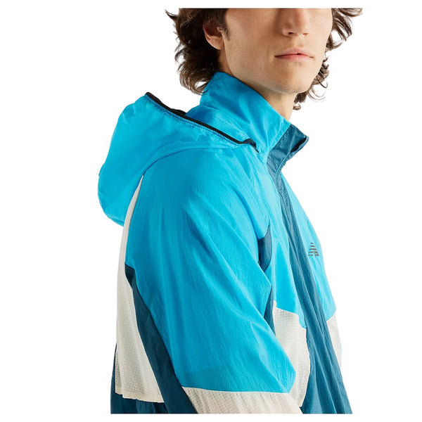 En mand med mellemlangt brunt hår har New Balance RC Jacket, Herre, Streamsong på – en blå og hvid vandafvisende jakke – og er afbilledet fra brystet og opefter, mens han står let vendt til siden foran en hvid baggrund.