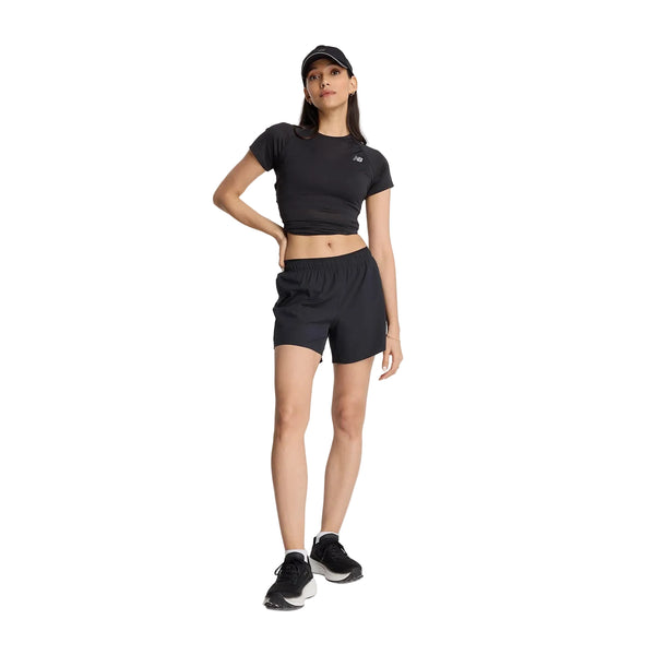 En person står foran en hvid baggrund iført en sort kasket, en sort kortærmet crop-top, et par New Balance Rc Short 5" Dame i sort og sorte sneakers, med den ene hånd på hoften.