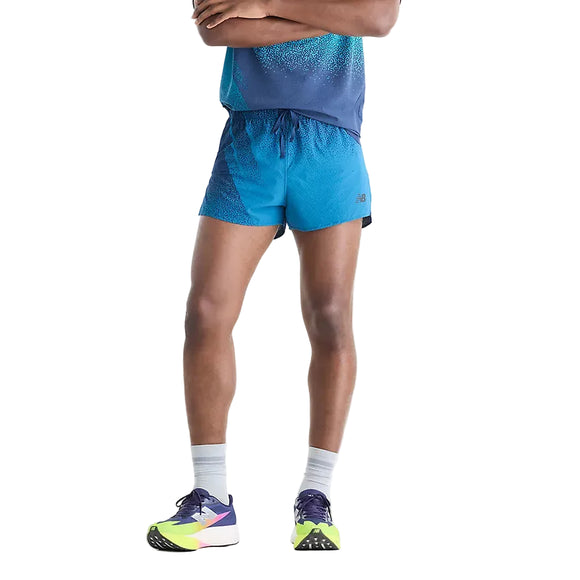En person står med armene over kors og er iført et blåt sportsoutfit med farveovergang, herunder et par New Balance Rc Ultra Light Printed Short 3″ til mænd i farven »Altitude«, grå sokker og neonfarvede løbesko. Billedet fokuserer på underkroppen foran en ensfarvet hvid baggrund.