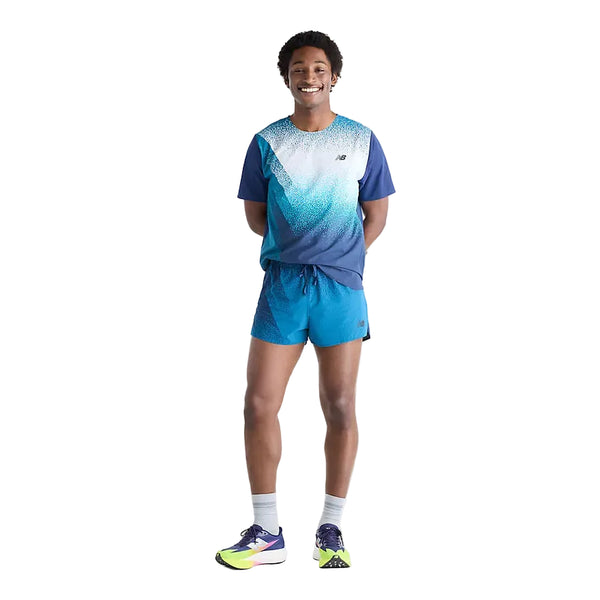 En mand står og smiler med hænderne bag ryggen. Han er iført en blå sports-T-shirt med farveovergang, New Balance Rc Ultra Light Printed Short 3″ Herre i farven Altitude, grå sokker og blå og gule løbesko foran en ensfarvet hvid baggrund.