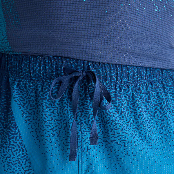 Nærbillede af en person, der bærer New Balance Rc Ultra Light Printed Short 3″, Herre, Altitude med elastisk linning og snøre, der kan bindes, over en blå skjorte med struktur.