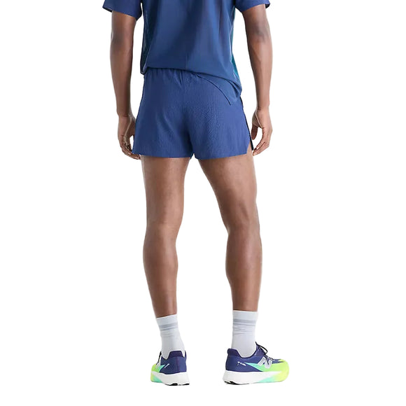 En person står med ryggen til kameraet iført et blåt træningssæt bestående af New Balance RC Ultra Light Printed Short 3″, Herre, Altitude, grå strømper samt blå og neongrønne New Balance-løbesko foran en ensfarvet hvid baggrund.