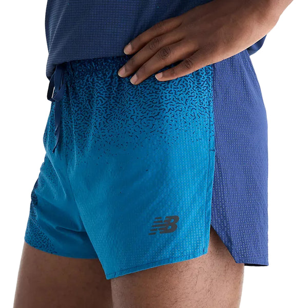En person iført blå New Balance Rc Ultra Light Printed Short 3″, Herre, Altitude-løbeshorts med et prikkemønster i farveovergang og slids i siden nederst. Højre hånd hviler på hoften, og en del af en matchende blå T-shirt kan ses øverst.