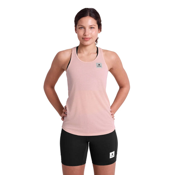 En kvinde står med ansigtet fremad og hænderne på hofterne, iført Saysky Reactive Flow Singlet, Dame, pink logo og sorte, tætsiddende shorts, klar til løbsdagen på en almindelig hvid baggrund.