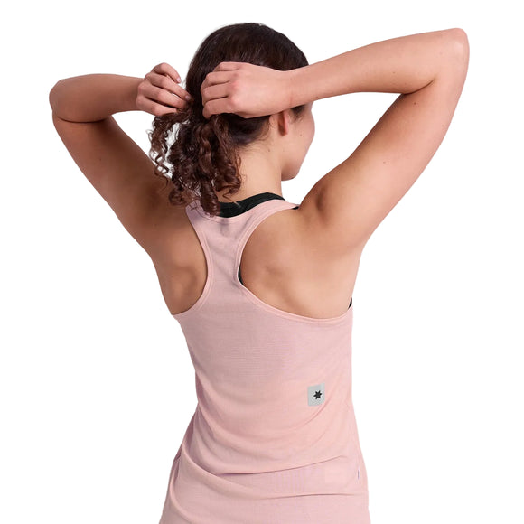 En person med krøllet brunt hår, set bagfra, er iført en Saysky Reactive Flow Singlet, Dame, Pink Logo og holder armene op for at binde håret. Toppen har et lille mærke nær den nederste del af ryggen.
