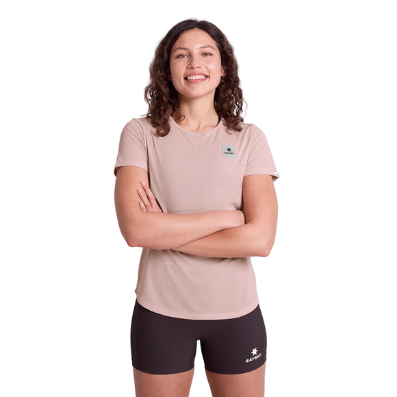 En person med krøllet brunt hår står på en hvid baggrund og smiler med armene over kors. De er iført en Saysky Reactive Flow T-shirt til kvinder med pink logo og sorte sportsshorts, begge med små logoer.