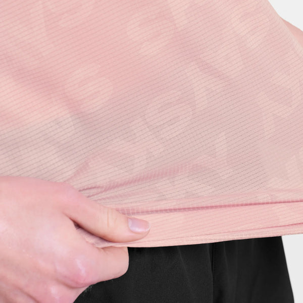 Nærbillede af en persons hånd, der holder kanten af en Saysky Reactive Flow T-shirt, Dame, Pink Logo, med svage diagonale linjer og et diskret mønster, sammen med sorte bukser.