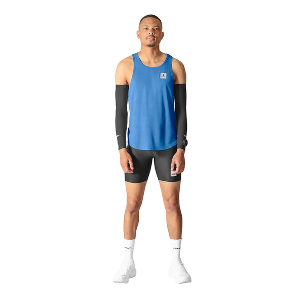 En person står med ansigtet fremad iført en blå Saysky Reactive Flow Singlet, Herre, Blue Aop-tanktop, der er designet til optimal svedtransport, sorte kompressionsærmer på begge arme, sorte shorts og hvide sokker samt hvide sportssko på en ensfarvet hvid baggrund.