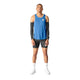 En person står med ansigtet fremad iført en blå Saysky Reactive Flow Singlet, Herre, Blue Aop-tanktop, der er designet til optimal svedtransport, sorte kompressionsærmer på begge arme, sorte shorts og hvide sokker samt hvide sportssko på en ensfarvet hvid baggrund.