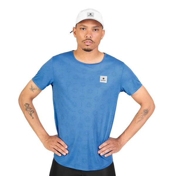 En mand iført en hvid kasket og en Saysky Reactive Flow T-shirt, Herre, Blue Aop, står med hænderne i siden. Han har tatoveringer på begge arme og et kortklippet skæg. Baggrunden er helt hvid.