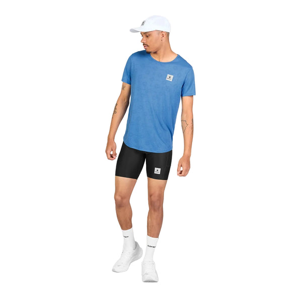 En mand iført en blå Saysky Reactive Flow T-shirt, Herre, Blue Aop, sorte sportsshorts, hvide sokker, hvide sportssko og en hvid baseballkasket står foran en hvid baggrund. Han kigger til venstre med armene afslappet ned langs siden. Den åndbare T-shirt fremhæver den karakteristiske komfort i Reactive Flow T-shirt-kollektionen.