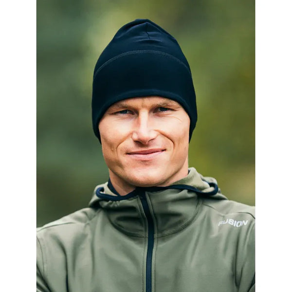 En person iført Fusion Recharge Beanie, Unisex, Black, og en olivengrøn jakke med Fusion-logo står udendørs og kigger ind i kameraet med et lille smil. Sløret grønt i baggrunden antyder udendørs sportsaktiviteter.